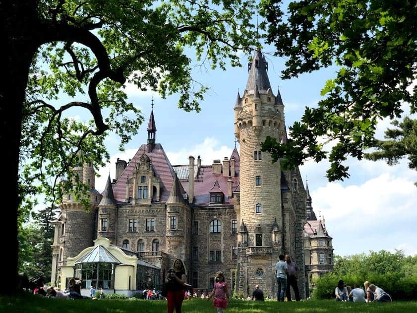 Large stone castle with turrets and red roofs, surrounded by people relaxing on a grassy lawn under leafy trees on a sunny day—a must-see castle for anyone interested in Poland tourism.