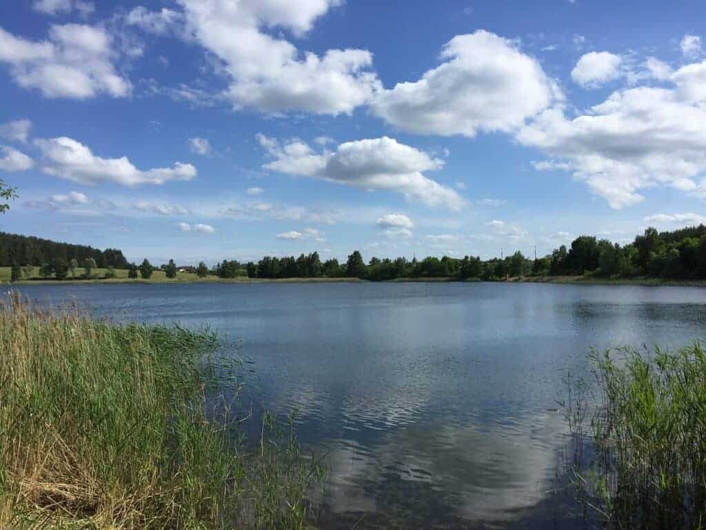 A calm lake in Mazury, Poland, surrounded by green reeds and trees under a partly cloudy blue sky—one of the best places to visit for tranquil travel moments.