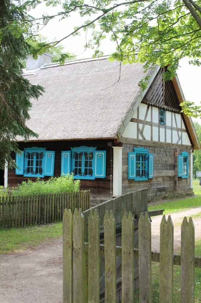 A traditional wooden house with a thatched roof, blue window shutters, and a picket fence, surrounded by greenery—perfect for experiencing the charm of Mazury on your Poland travel adventure.