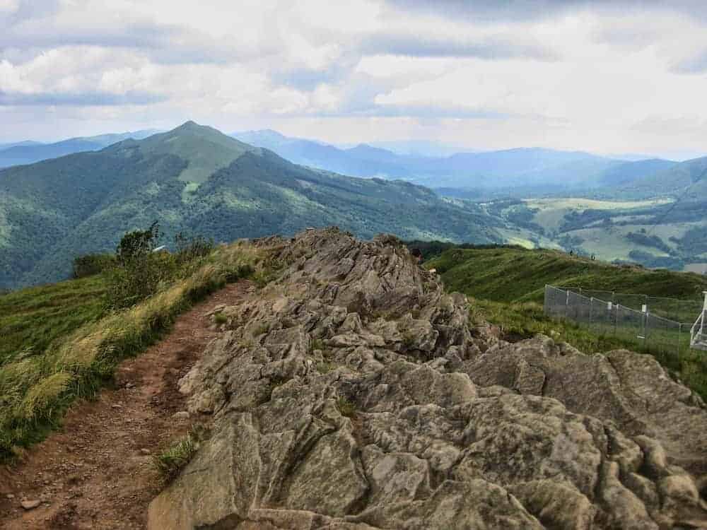 A rocky trail runs along a ridge with grassy hills and distant mountains under a cloudy sky. A metal fence is visible on the right side.