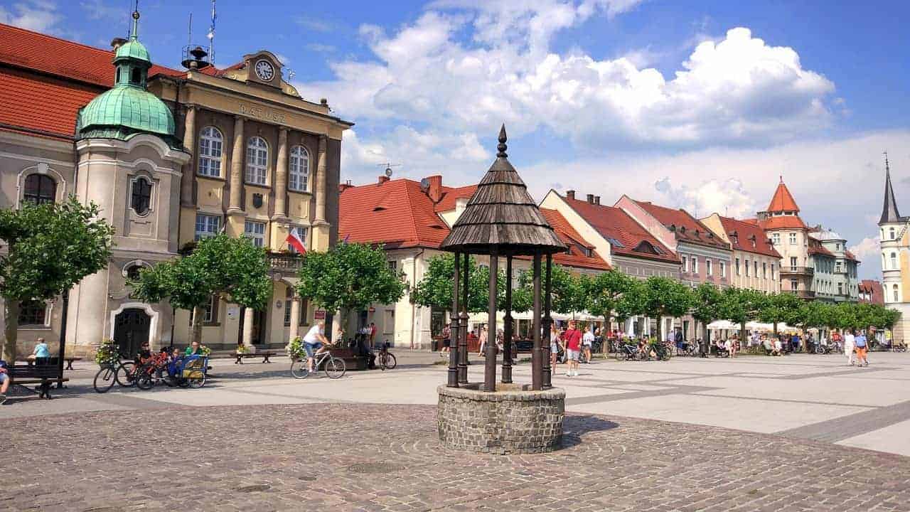 A stone well stands in the center of a cobblestone town square in Slaskie, Poland, surrounded by historic buildings with red roofs, trees, and people walking or cycling—one of the best destinations to visit.