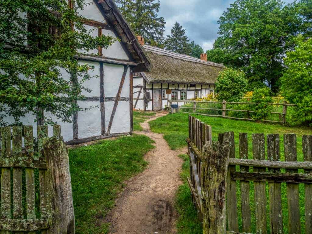 A dirt path leads through a wooden gate to traditional half-timbered houses with thatched roofs, surrounded by green trees and grass—a breathtaking scene reminiscent of Poland’s charming National Parks.