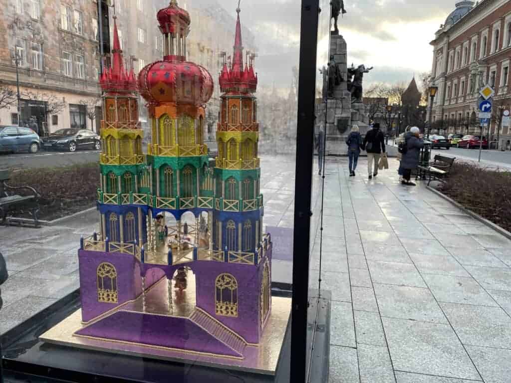 A colorful, miniature model of a castle is displayed in a glass case on a city sidewalk, inviting passersby to experience the charm of Krakow travel amid historic buildings—even during winter in Krakow.