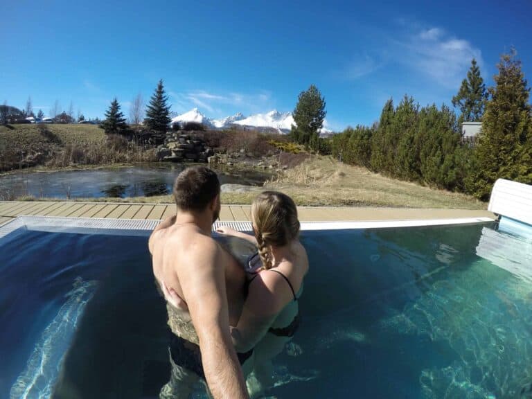 Two people stand together in an outdoor thermal bath in Zakopane, surrounded by trees and grass, with snow-capped mountains visible in the distance under a clear blue sky.
