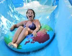 A woman in a swimsuit rides an inflatable tube down a blue water slide at one of Poland’s exciting water parks, smiling with her mouth open.