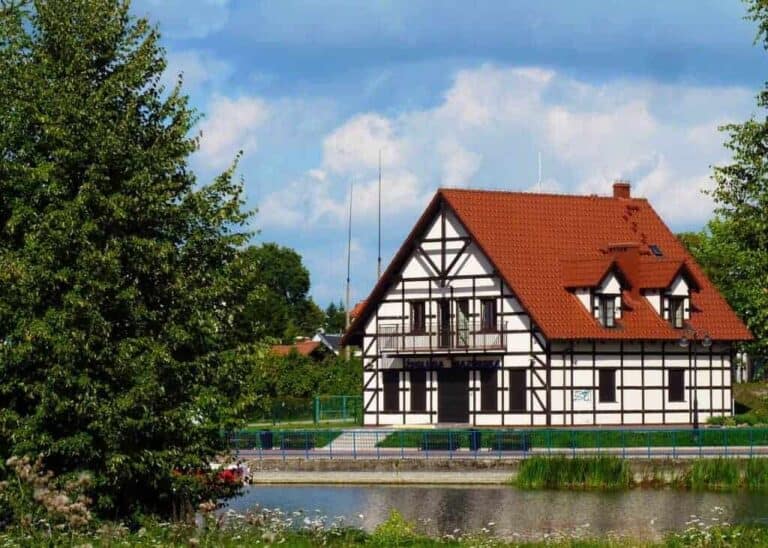 A white half-timbered house with a red roof stands by a pond, surrounded by trees and greenery under a partly cloudy sky—an idyllic scene that makes Mazury, Poland one of the best places to visit for travel enthusiasts.
