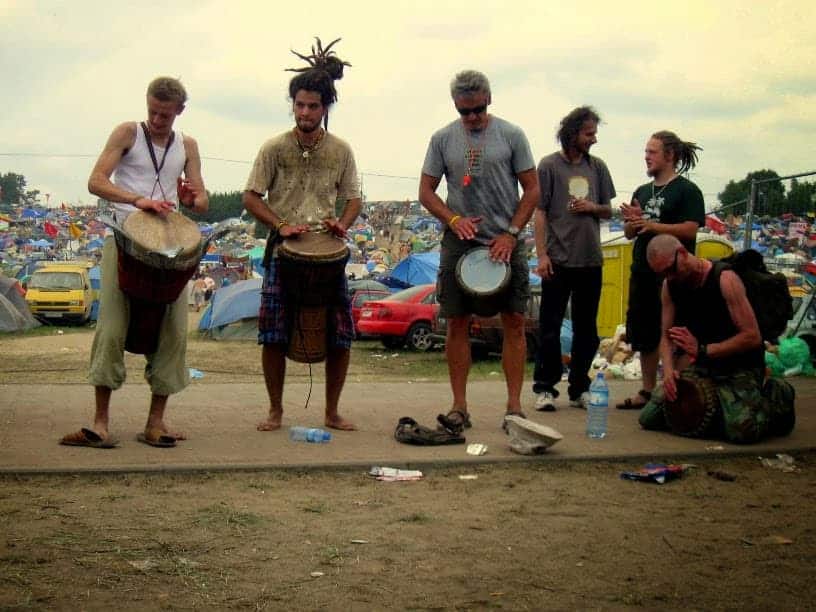Five people play hand drums outdoors near tents and parked cars, with many people and camping gear visible in the background—a lively scene capturing the spirit of summer in Poland.