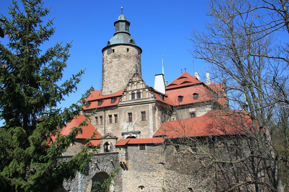 A large stone castle with a round tower, red-tiled roofs, and arched windows, surrounded by trees under a clear blue sky—one of the must-see castles in Poland for any Poland tourism enthusiast.