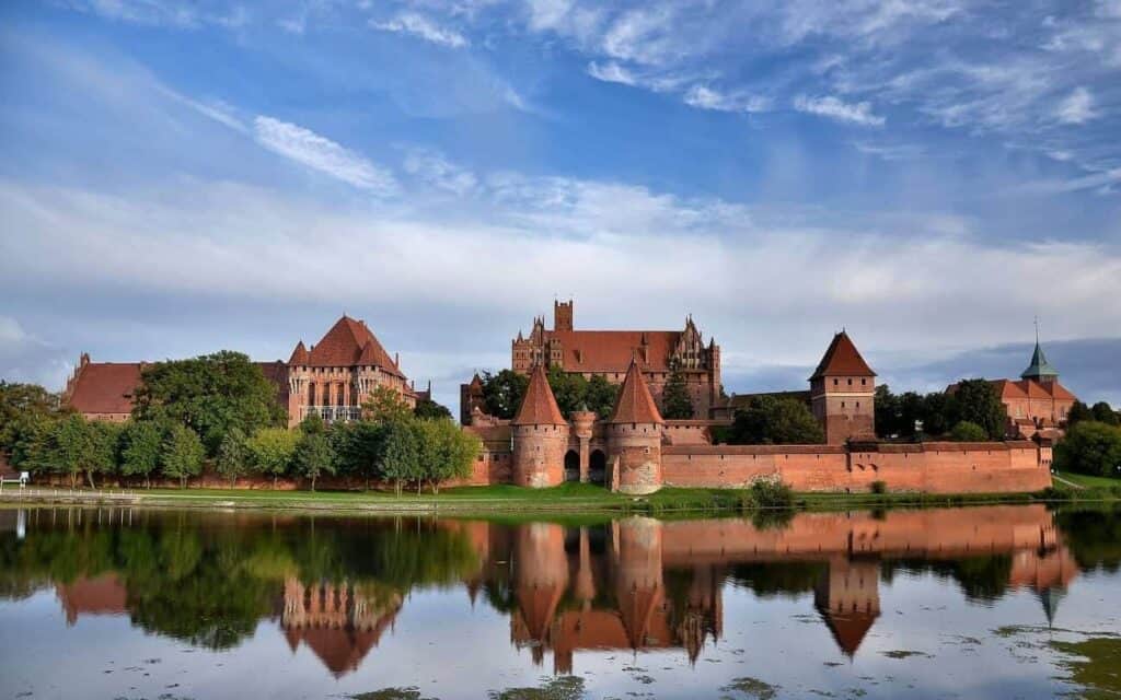 Large brick castle complex with towers and walls, surrounded by greenery and reflected in a calm body of water under a blue sky with scattered clouds—a true highlight among must-see castles for Poland tourism.