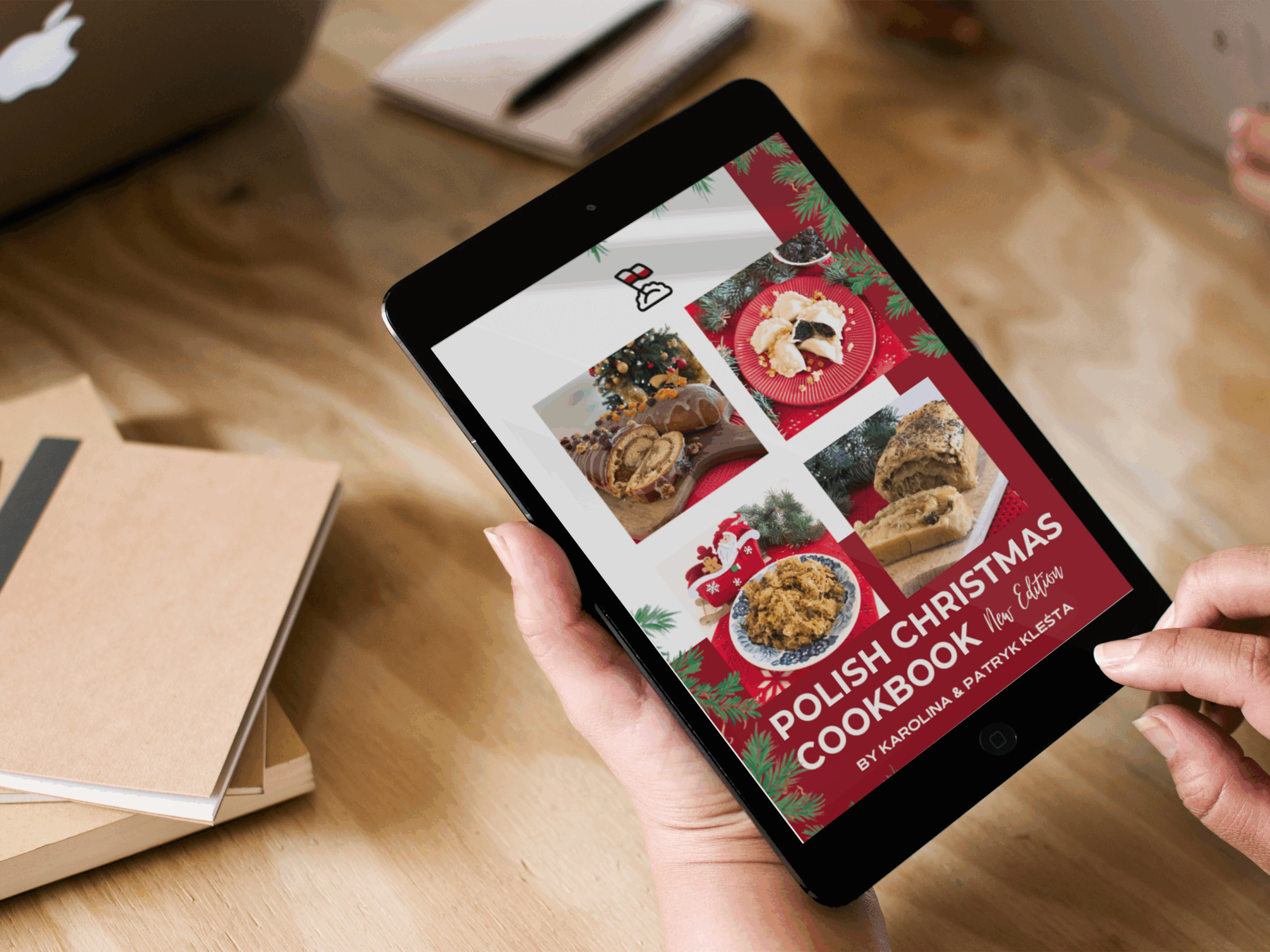 A person holds a tablet displaying the cover of a "Polish Christmas Cookbook," part of a festive Christmas Kit, featuring food photos and holiday decorations on a wooden table with notebooks.