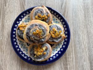 Five iced poppy seed rolls topped with chopped candied orange sit on a decorative blue and white plate on a wooden surface.